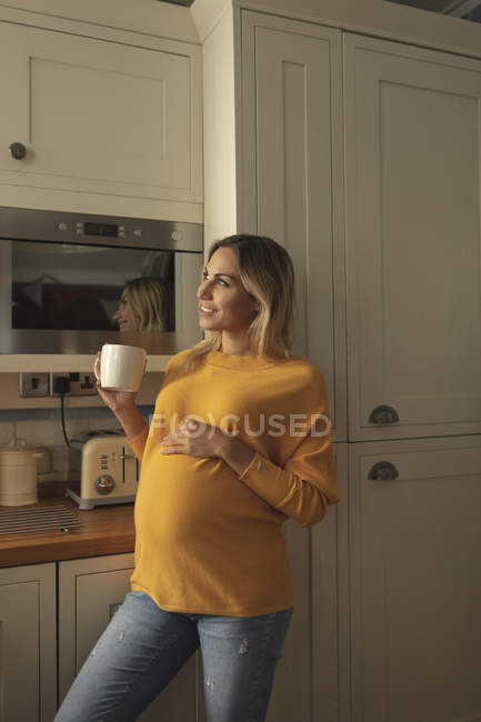 Усміхнена вагітна жінка з чашкою кави стоїть на кухні — стокове фото