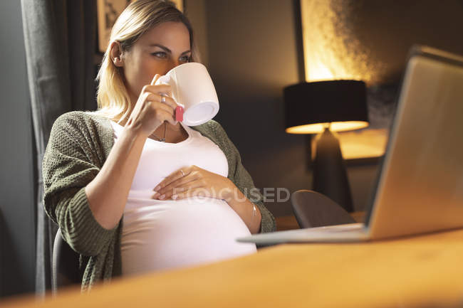Mujer embarazada bebiendo café mientras usa el ordenador portátil en casa - foto de stock