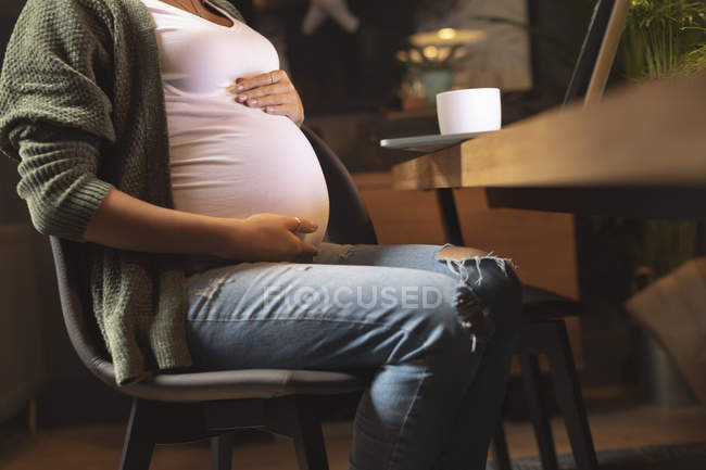 Sección media de la mujer embarazada tocándose el vientre mientras usa la computadora portátil - foto de stock