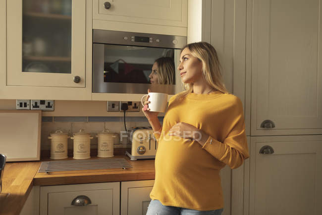 Donna incinta premurosa con tazza di caffè in piedi in cucina — Foto stock