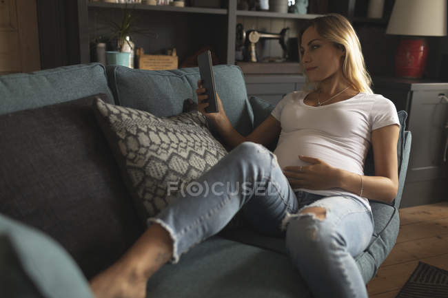 Беременная женщина читает цифровой планшет на диване дома — стоковое фото