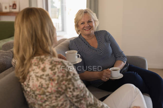 Madre e hija interactuando entre sí mientras toman café en la sala de estar en casa - foto de stock