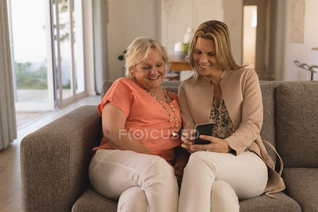 Madre e hija usando el teléfono móvil en la sala de estar en casa - foto de stock