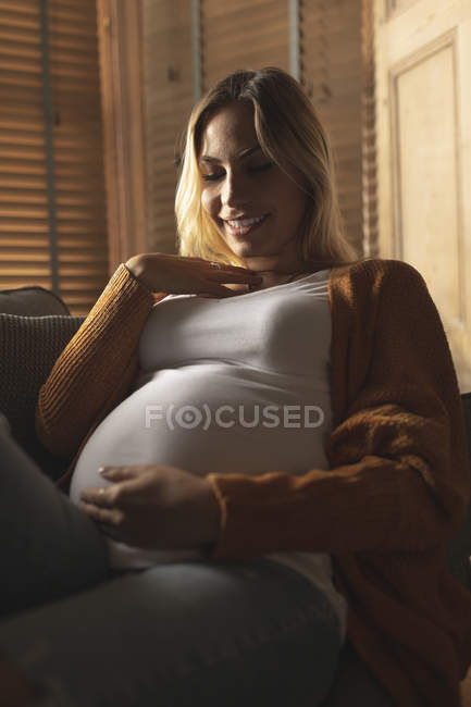 Усміхнена вагітна жінка сидить на дивані, торкаючись її живота — стокове фото