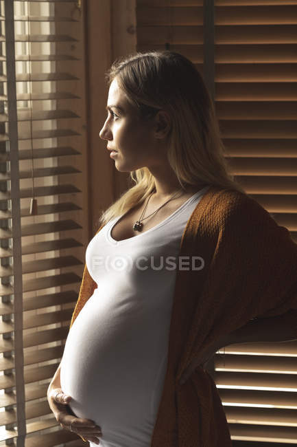 Thoughtful pregnant woman looking through window blinds — Stock Photo