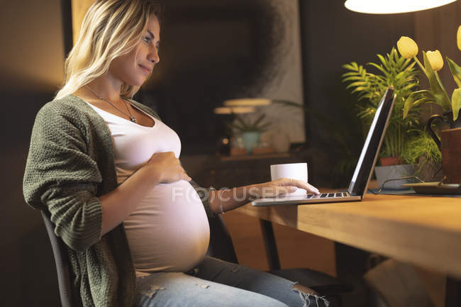 Беременная женщина трогает живот и пользуется ноутбуком дома — стоковое фото