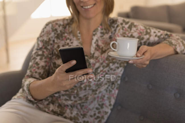 Sección media de la mujer que usa el teléfono móvil mientras toma café en casa - foto de stock