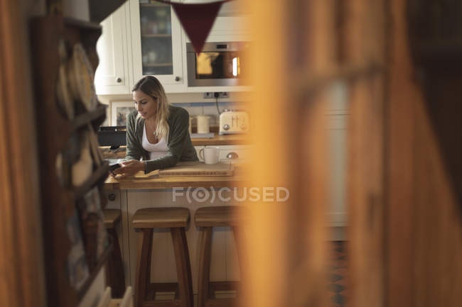 Mujer embarazada mensajes de texto en el teléfono en la cocina en casa - foto de stock