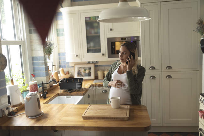 Mujer embarazada hablando por teléfono en la cocina en casa - foto de stock