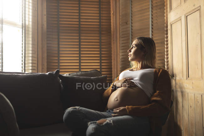 Беременная женщина дотрагивается до живота, глядя в окно дома. — стоковое фото