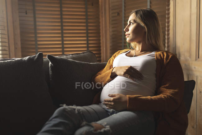 Mujer embarazada sentada en el sofá mirando a través de la ventana en casa - foto de stock