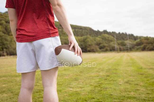 Средняя секция регбиста, стоящего с мячом для регби на поле — стоковое фото