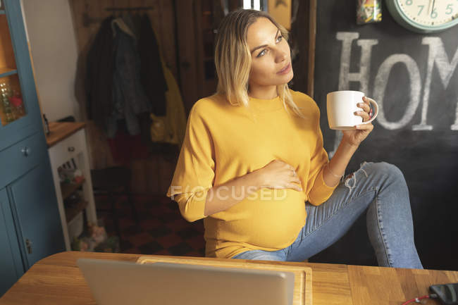 Pregnant woman with coffee cup looking out of the window at home — Stock Photo