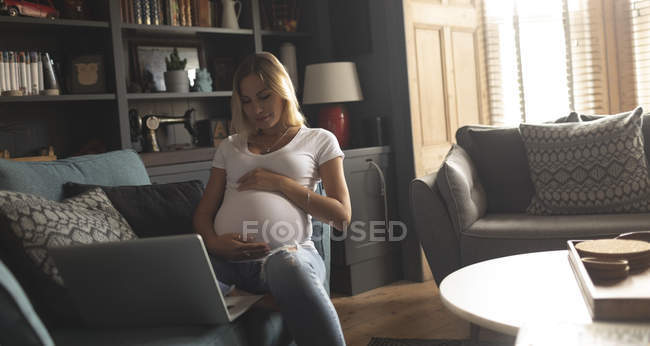 Femme enceinte assise sur un canapé qui touche son ventre à la maison — Photo de stock