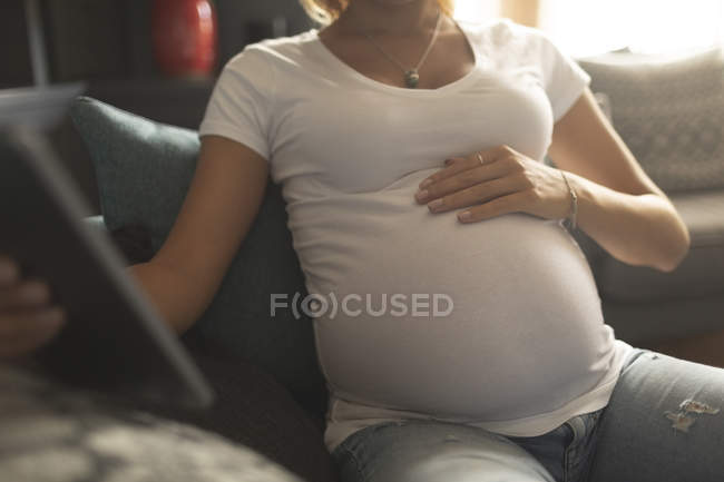 Partie médiane de la femme enceinte utilisant une tablette numérique sur le canapé — Photo de stock