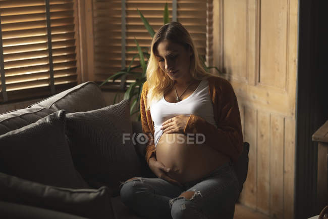 Mulher grávida sentada no sofá tocando sua barriga em casa — Fotografia de Stock