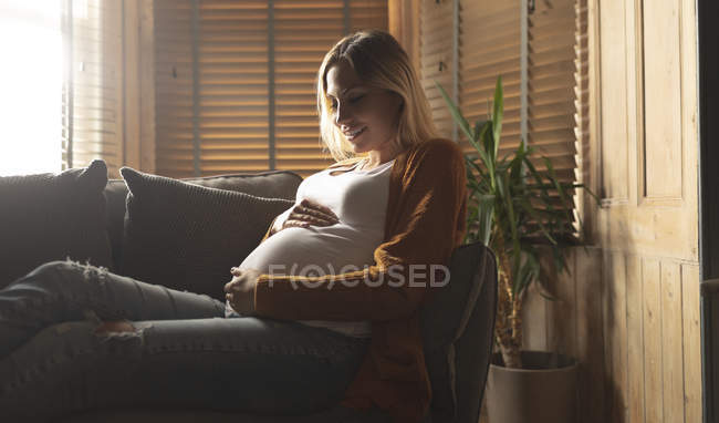 Sorridente donna incinta seduta sul divano toccare la pancia — Foto stock