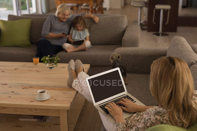 Femme utilisant un ordinateur portable tandis que grand-mère et petite-fille utilisant un téléphone mobile et une tablette numérique à la maison — Photo de stock