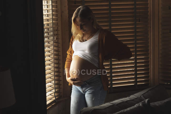 Беременная женщина прикасается к животу у окна дома — стоковое фото