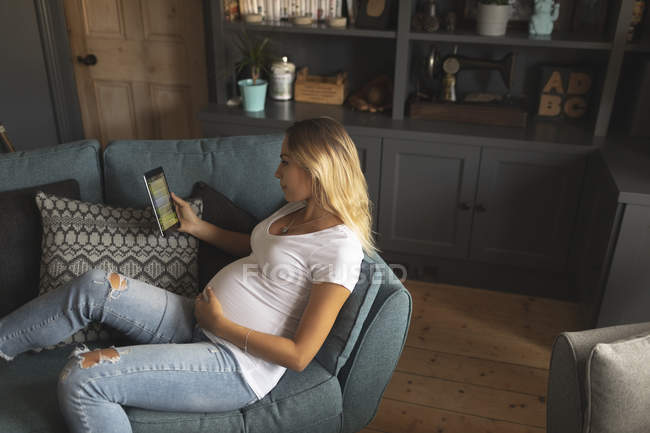 Беременная женщина читает цифровой планшет на диване дома — стоковое фото