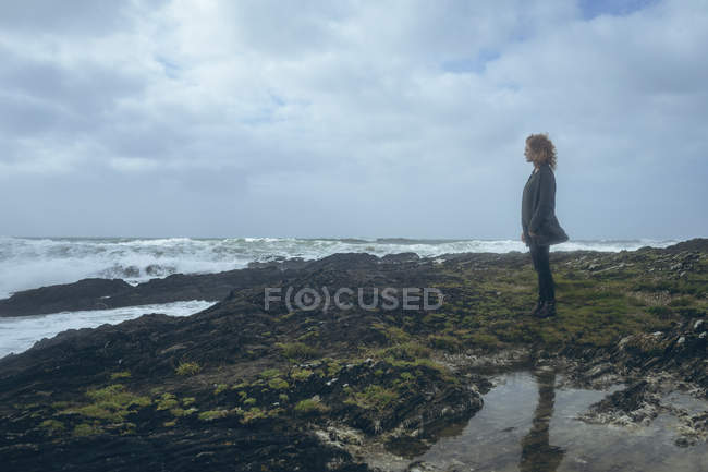 Pensativo pelirroja mujer de pie en la playa en las rocas . - foto de stock