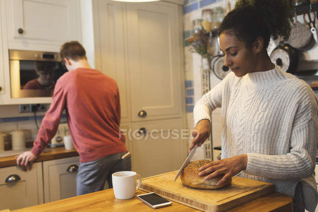 Woman cutting loaf of bread in kitchen at home — Stock Photo