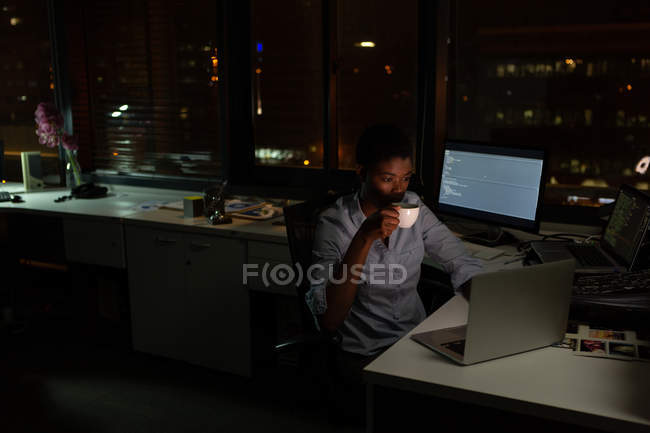 Esecutivo femminile che prende il caffè mentre usa il computer portatile in ufficio di notte — Foto stock
