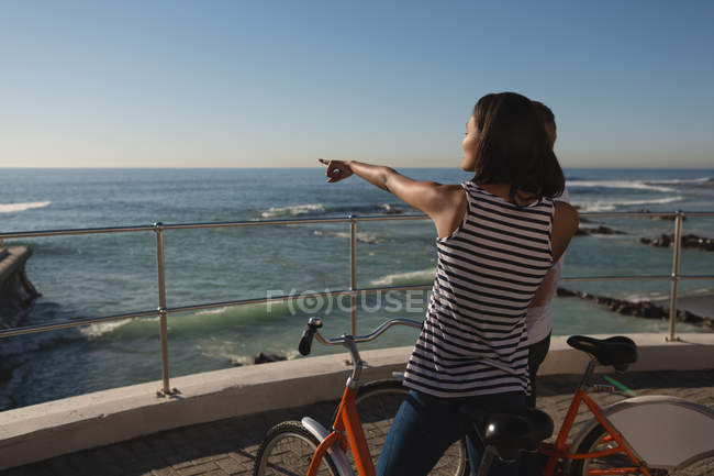 Young woman with bicycle standing on promenade and pointing to sea near beach — Stock Photo