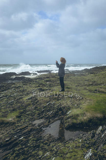 Side view of the redhead woman taking photo with mobile phone in the beach . — стоковое фото