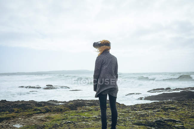 Vue arrière de la femme rousse en utilisant un casque de réalité virtuelle dans une plage venteuse . — Photo de stock