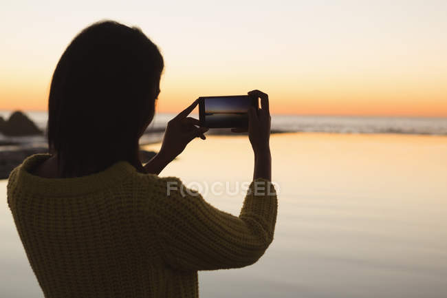 Donna che scatta foto con cellulare in spiaggia durante il tramonto — Foto stock