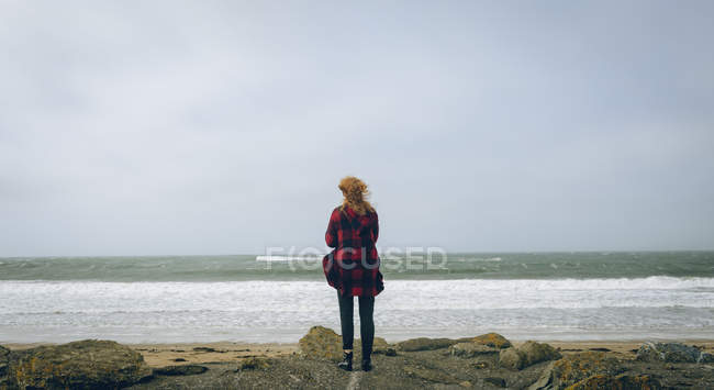 Вид ззаду руда жінка стоїть на вітряному пляжі . — стокове фото