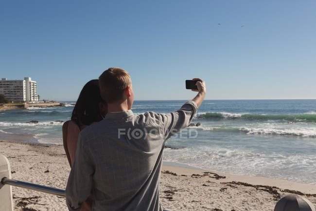 Vista posteriore di coppia che prende selfie sul lungomare vicino alla spiaggia — Foto stock