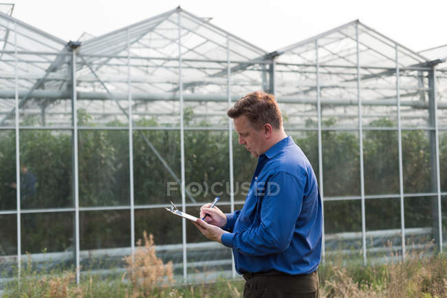 Concentrated man writing on clipboard in greenhouse — Stock Photo