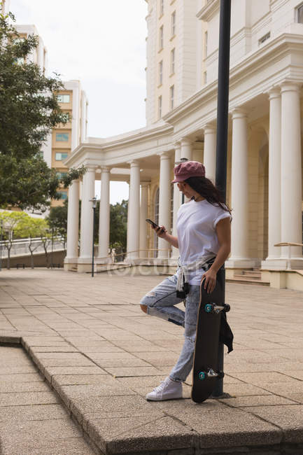 Vista lateral do skatista feminino usando telefone celular na cidade — Fotografia de Stock