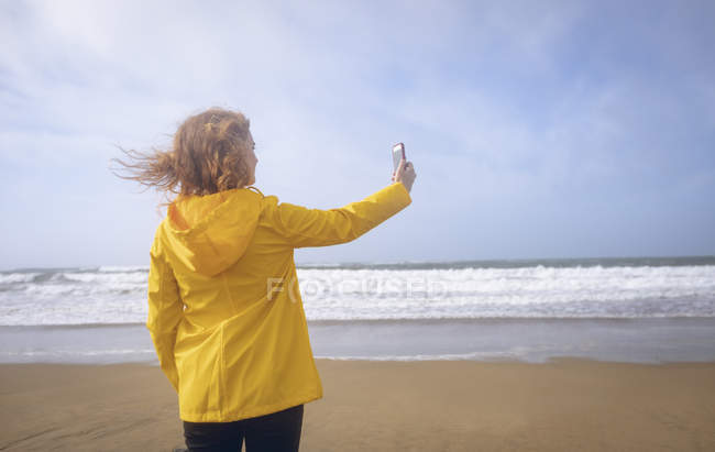 Vista posteriore della donna rossa che prende selfie con il telefono cellulare in spiaggia . — Foto stock