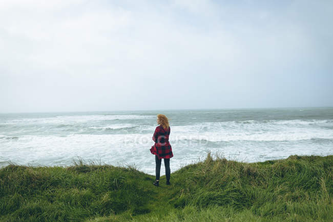 Vista trasera de mujer pelirroja de pie en la playa cubierta de hierba . - foto de stock