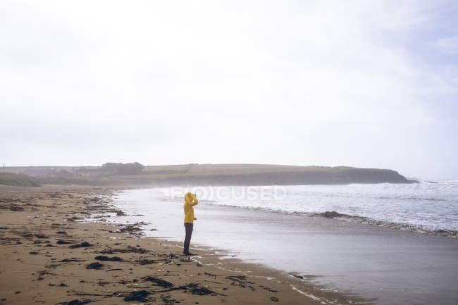 Vista lateral da mulher de casaco amarelo em pé na praia . — Fotografia de Stock