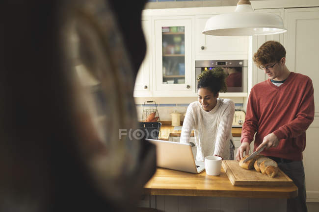 Mujer usando el ordenador portátil, mientras que el hombre cortar pan en la cocina en casa - foto de stock
