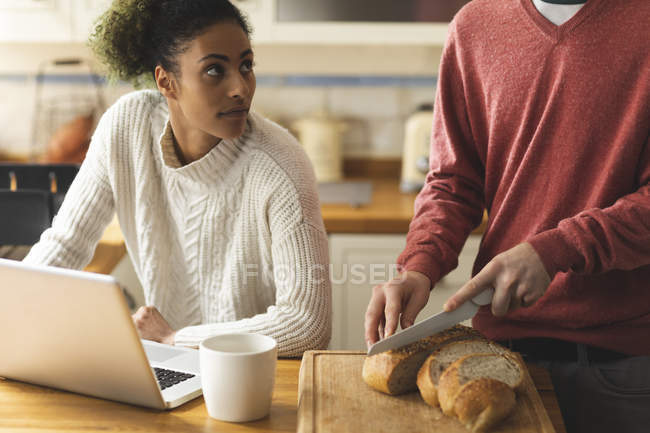 Woman using laptop while man cutting loaf of bread in kitchen at home — Stock Photo