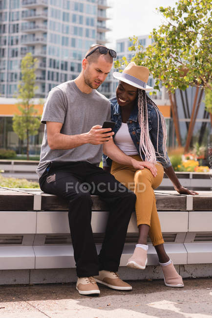 Lächelndes Paar sitzt auf Bank und benutzt Handy in der Stadt — Stockfoto