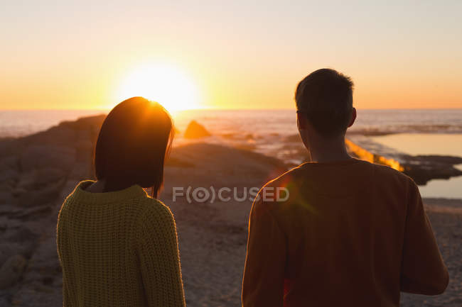 Вид сзади пары, стоящей на пляже во время заката — стоковое фото