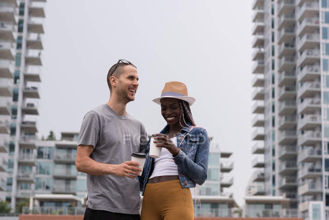 Jovem casal feliz com xícaras de café em pé contra edifícios da cidade — Fotografia de Stock