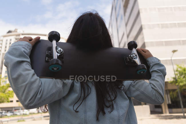 Вид сзади на скейтбордистку, несущую скейтборд в городе — стоковое фото