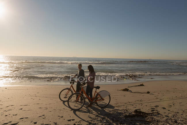 Pareja con bicicletas caminando en la playa a la luz del sol - foto de stock