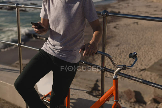 Середина чоловіка з велосипедом, використовуючи мобільний телефон біля перил на пляжі — стокове фото