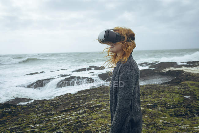 Mulher ruiva usando fone de ouvido realidade virtual na praia . — Fotografia de Stock