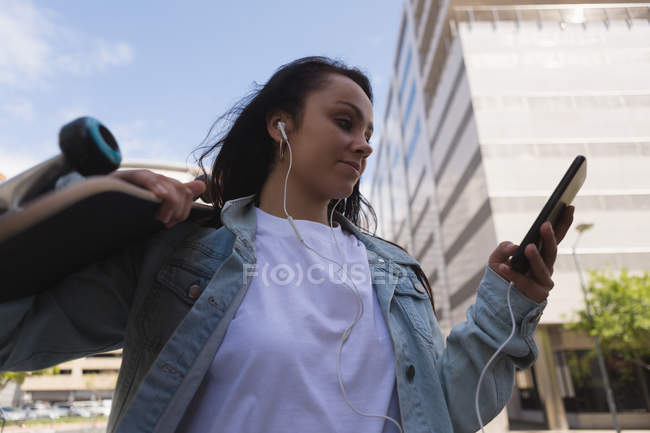 Низкий угол обзора женского скейтбординга, слушающего музыку на мобильном телефоне — стоковое фото