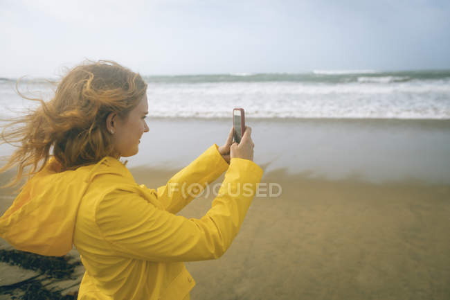 Femme rousse prenant des photos avec téléphone portable à la plage . — Photo de stock