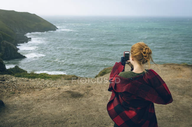 Vista trasera de la mujer pelirroja tomando fotos con teléfono móvil en la playa . - foto de stock